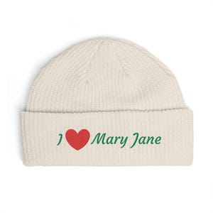 Op de afbeelding: Crèmekleurige beanie met een omgeslagen rand. De hoed heeft de tekst "I ❤️ Mary Jane" in groene letters met een rood hartsymbool. De hoed is gemaakt van een geribbelde gebreide stof.