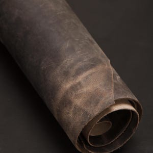 Puede incluir: Material de cuero marrón oscuro enrollado. El cuero tiene un aspecto envejecido con variaciones de color y textura. El rollo está bien enrollado, revelando las capas internas. El fondo es de color oscuro sólido.