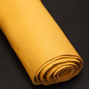 Puede incluir: Un trozo de cuero amarillo enrollado sobre un fondo negro. El cuero tiene una superficie texturizada y parece suave. Los bordes del rollo son visibles, mostrando las capas del material. El color es amarillo mostaza.