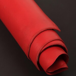 May include: A tightly rolled sheet of vibrant red leather. The material displays a smooth, matte surface texture with visible edges showing the thickness of the hide. The roll is positioned diagonally, highlighting the consistent red color throughout.