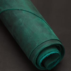 Könnte beinhalten: Ein aufgerolltes Stück smaragdgrünes Leder. Das Leder hat ein abgenutztes, strukturiertes Aussehen mit Farbvariationen. Die Kanten der Rolle zeigen einen helleren Türkiston. Der Hintergrund ist eine dunkle, einfarbige Farbe.