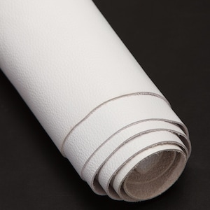 Puede incluir: Una hoja de cuero blanco enrollada, mostrando su superficie texturizada y las capas del material. Los bordes revelan un reverso más claro y sin terminar, sobre un fondo oscuro.