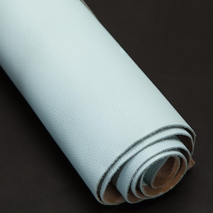 Puede incluir: Un trozo de cuero azul claro enrollado. El cuero tiene una superficie texturizada y una parte inferior marrón. El rollo está colocado en diagonal sobre un fondo oscuro, mostrando el color y la textura del material.