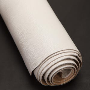 Puede incluir: Un rollo de cuero blanco roto enrollado. El cuero tiene una superficie texturizada y está enrollado firmemente, revelando los bordes del material. El fondo es negro sólido, proporcionando contraste con el cuero claro.