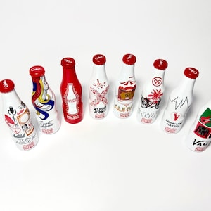 May include: Eight Coca-Cola aluminum bottles arranged in a semi-circle, each featuring unique graphic designs, red caps, and branding. The bottles display various illustrations, text, and logos, including names like Tony Aguilar and Abraham Mateo.