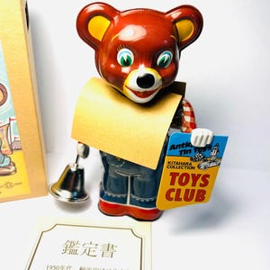 Può includere: Un orsetto giocattolo vintage in latta con corpo rosso, orecchie gialle e tuta blu. L'orso tiene in mano un cartello con la scritta "TOYS CLUB" e una campana d'argento. Sono visibili anche un certificato e una scatola.