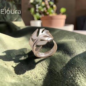 Può includere: Anello color argento a forma di foglia tropicale, con dettagli delle venature. L'anello è su uno sfondo verde, con la parola "Eloura". Il design ricorda una foglia di Monstera.