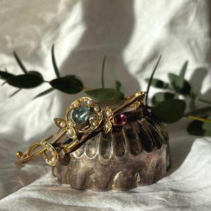 Puede incluir: Una tiara dorada con un diseño ornamentado, con una piedra preciosa central de color turquesa y pequeñas perlas. También se ve una tiara más pequeña con una piedra preciosa rosa. La joya descansa sobre un recipiente decorativo plateado.