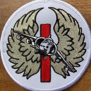 Könnte beinhalten: Runder, gestickter Aufnäher mit weißem Rand. Er zeigt ein stilisiertes Flugzeug mit beigefarbenen Flügeln und einem roten vertikalen Streifen. Das Flugzeug ist schwarz und weiß. Der Aufnäher ist aus Stoff.