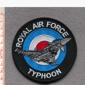 May include: A circular patch with the text "ROYAL AIR FORCE TYPHOON" in white. The patch features a jet aircraft design in grey and blue, set against a blue and red target background. The patch is approximately 10 cm in diameter.