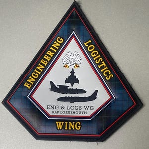May include: A diamond-shaped patch with a tartan pattern border. The patch features the words "ENGINEERING" and "LOGISTICS" in gold lettering. The center has a white background with a crest and two silhouettes of airplanes. The text "ENG & LOGS WG RAF LOSSIEMOUTH WING" is also present.