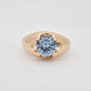 Op de afbeelding: Een gouden ring met een enkele, ronde, lichtblauwe gefacetteerde edelsteen in een gladde, gepolijste gouden band. De zetting heeft een ontwerp met vier pootjes die de centrale steen vasthouden, wat zorgt voor een strak en klassiek uiterlijk.