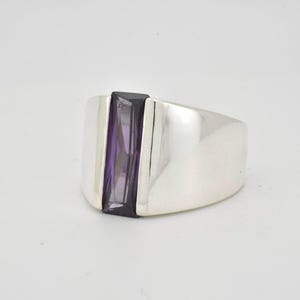 May include: A silver ring featuring a rectangular purple gemstone set within a black frame. The ring has a broad band and a polished finish, displaying a contemporary design. The gemstone is a vivid purple colour.