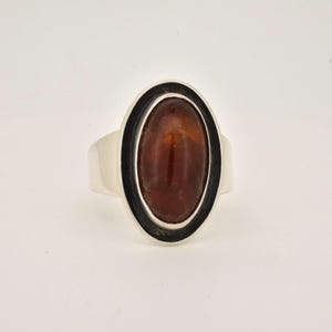 Op de afbeelding: Een zilveren ring met een ovale, amberkleurige steen. De steen is gezet in een zwarte rand, en de ringband is van gepolijst zilver. De ring wordt getoond tegen een witte achtergrond.