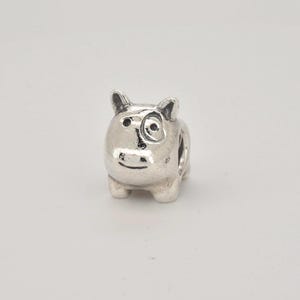 Może przedstawiać: Srebrzysty metalowy koralik typu charms w kształcie małego, stylizowanego psa z zaokrąglonym tułowiem, spiczastymi uszami i wyraźną ciemną plamką nad jednym okiem. Koralik ma gładkie, polerowane wykończenie z wgłębionymi czarnymi detalami.