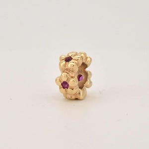 May include: A gold-colored charm with a floral design. The charm features three flower clusters, each with a small, round, red gemstone in the center. The charm is likely made of gold and is designed to be worn as jewelry.