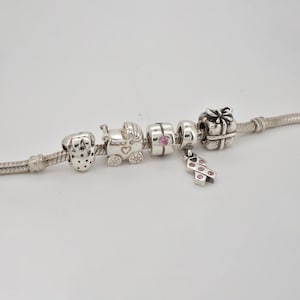 May include: A silver charm bracelet featuring various charms. The charms include a dog, a baby carriage, a charm with a pink gem, a gift box, and a pink ribbon. The bracelet is set against a white backdrop.