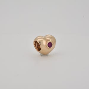 May include: A gold-coloured heart-shaped charm with a small, round, red gemstone. The charm has a hollowed-out section on one side. The charm is set against a plain white background. The charm is likely made of metal.