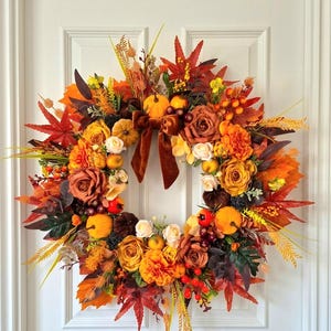 May include: A circular autumn wreath featuring a mix of orange, red, and yellow faux maple leaves, roses, and marigolds. The arrangement includes small decorative pumpkins, berries, and wheat stalks, accented by a brown velvet ribbon bow at the top.