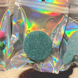 May include: Three teal-colored, decorative soap bars in heart, circle, and bunny shapes, each packaged in a holographic bag. Each soap has a silver wire loop and is accompanied by colorful, round beads in pink, green, and yellow.