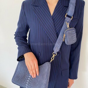 May include: A blue snakeskin-textured shoulder bag with a matching wide crossbody strap and a small detachable pouch. The bag and strap feature gold-tone hardware. The set is worn over a navy blue pinstriped blazer with a double-breasted front.
