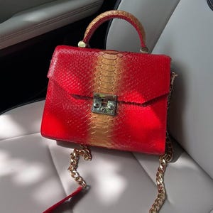 Może przedstawiać: Czerwona i złota torebka ze skóry węża z górnym uchwytem i złotym paskiem na łańcuszku. Torba ma prostokątny kształt z klapką i srebrnym zapięciem. Uchwyt i góra torby są złote.