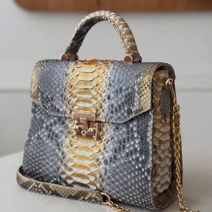 May include: A structured handbag with a snakeskin pattern in grey, yellow, and white. The bag features a top handle, a gold-tone clasp, and a gold chain strap. The bag is sitting on a white surface.
