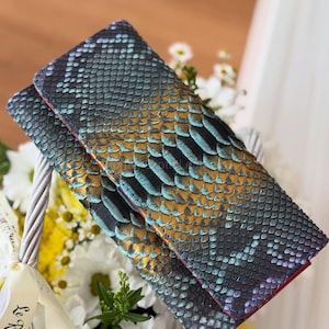 Puede incluir: Una cartera plegable con un patrón de piel de serpiente en tonos turquesa, dorado y negro. La cartera tiene un interior rojo y se exhibe sobre un lecho de flores blancas y amarillas. Las escamas son visibles y detalladas.