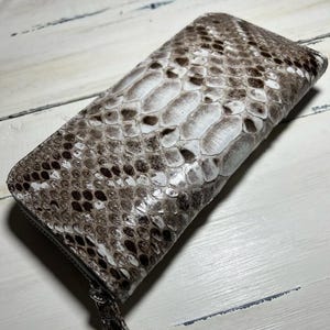 May include: A rectangular wallet with a zip closure, featuring a snakeskin pattern in shades of brown, white, and black. The wallet's texture is visible, showcasing the scales. The zip pull is silver.