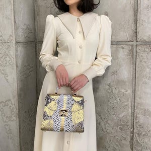 May include: A cream-coloured dress with a large collar and long sleeves. The dress is buttoned down the front. A square handbag with a gold clasp and a snakeskin pattern in gold, white, and grey is held in front of the dress.