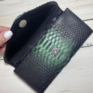 Puede incluir: Clutch negro y verde con textura de piel de serpiente y cierre magn&eacute;tico. El interior es negro con un bolsillo con cremallera. El clutch est&aacute; abierto, revelando el interior y el cierre magn&eacute;tico.