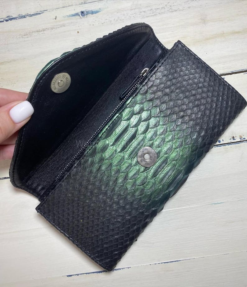 Puede incluir: Clutch negro y verde con textura de piel de serpiente y cierre magn&eacute;tico. El interior es negro con un bolsillo con cremallera. El clutch est&aacute; abierto, revelando el interior y el cierre magn&eacute;tico.