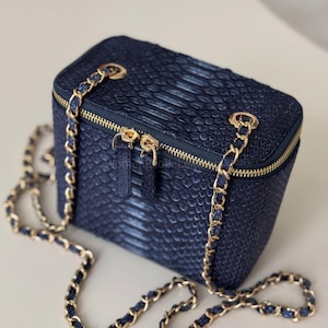 Op de afbeelding: Een marineblauwe rechthoekige handtas met een textuur van slangenleer. De tas heeft een goudkleurige ritssluiting en een kettingriem. De kettingriem is ongeveer 122 cm lang.