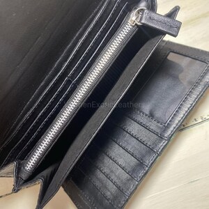 Puede incluir: Cartera de cuero negro abierta con m&uacute;ltiples compartimentos, incluyendo ranuras para tarjetas y una secci&oacute;n con cremallera. La cartera est&aacute; abierta, revelando el dise&ntilde;o interior. La cremallera es plateada.