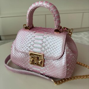 May include: A small, pink handbag with a top handle and a gold chain strap. The bag has a textured, scaly pattern and a gold-coloured clasp. The handle and strap are also pink.