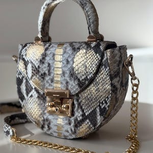 May include: A small, round handbag with a snakeskin pattern in shades of grey, black, and gold. The bag features a top handle, a gold chain strap, and a gold clasp closure. The bag is a stylish accessory.