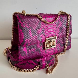 May include: A pink and black snakeskin-effect handbag with a gold chain strap and clasp. The bag is rectangular with a flap closure and a gold chain strap for shoulder or cross-body wear. The bag is made of a snakeskin-effect material.