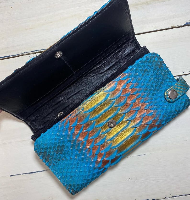 K&ouml;nnte beinhalten: Ge&ouml;ffnete Geldb&ouml;rse aus blauem, goldenem und orangefarbenem Schlangenleder mit schwarzem Innenraum. Sie hat einen Druckknopfverschluss und einen silbernen Knopf. Das Schlangenmuster ist auf der Au&szlig;enseite sichtbar.