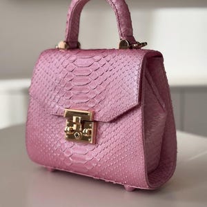 May include: A pink handbag with a snakeskin texture. It features a top handle, a flap closure, and gold-coloured hardware. The bag is displayed on a white surface.