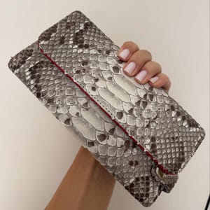 Może przedstawiać: Szary i biały portfel ze wzorem skóry węża z czerwonym akcentem. Prostokątny portfel ma zapięcie na guzik. Wzór skóry węża jest szczegółowy, z odcieniami szarości i bieli. Portfel ma około 20 cm długości.