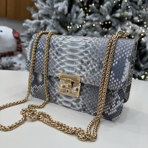 Op de afbeelding: Een grijze en witte handtas met slangenpatroon, een gouden kettingriem en een gouden sluiting. De tas is rechthoekig met een klepsluiting. De achtergrond toont een besneeuwde kerstboom.