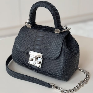 Może przedstawiać: Mała, czarna torebka z teksturowanym wzorem przypominającym skórę węża. Torebka posiada górny uchwyt, srebrny łańcuszek i srebrne zapięcie. Torebka stoi na białej powierzchni.