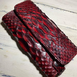 May include: A red python skin clutch bag with a textured, scaled pattern. The bag has a fold-over flap and a silver-toned snap closure. The colour is a deep, rich red with darker accents.