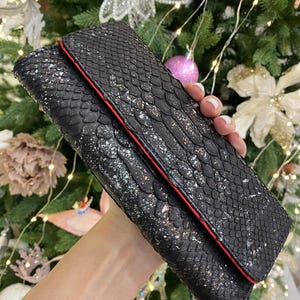 May include: A black clutch bag with a snakeskin effect and a red trim. The bag has a fold-over flap and is decorated with silver flecks. The rectangular bag appears to be made of leather.