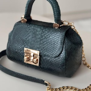 Może przedstawiać: Ciemnozielona torebka z teksturowanym wzorem przypominającym skórę węża. Torebka posiada górny uchwyt, złoty łańcuszek i złote zapięcie. Torebka ma szerokość około 20 cm.