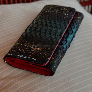 May include: A black leather wallet with a textured, scale-like pattern and gold speckles. The interior and trim are red. The wallet is closed and resting on a striped, beige surface.