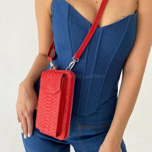 May include: A red snakeskin-effect crossbody phone bag with a matching strap and silver-tone hardware. The bag has a zip closure and a flap. The bag is shown against a blue denim background.