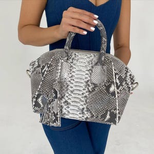 Op de afbeelding: Een grijze en witte handtas met slangenpatroon, met een handvat aan de bovenkant en een kwast. De tas heeft een afgeronde vorm en een ritssluiting. De tas wordt vastgehouden door een persoon die een blauwe spijkerstof top draagt.