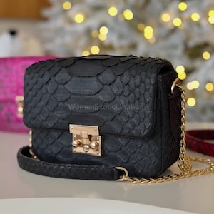 Op de afbeelding: Zwarte crossbodytas met een gouden ketting en sluiting. De tas heeft een klepsluiting en een schubachtige textuur. De tekst "WomenExoticLeathers" is zichtbaar op de zijkant. Een stijlvol accessoire.