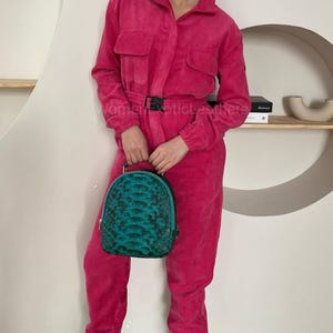 Op de afbeelding: Een felroze jumpsuit met lange mouwen, een kraag, twee borstzakken en een zwarte gespriem in de taille. De jumpsuit wordt gecombineerd met een afgeronde rugzak met een gestructureerd, schubachtig patroon in tinten van blauwgroen en zwart.